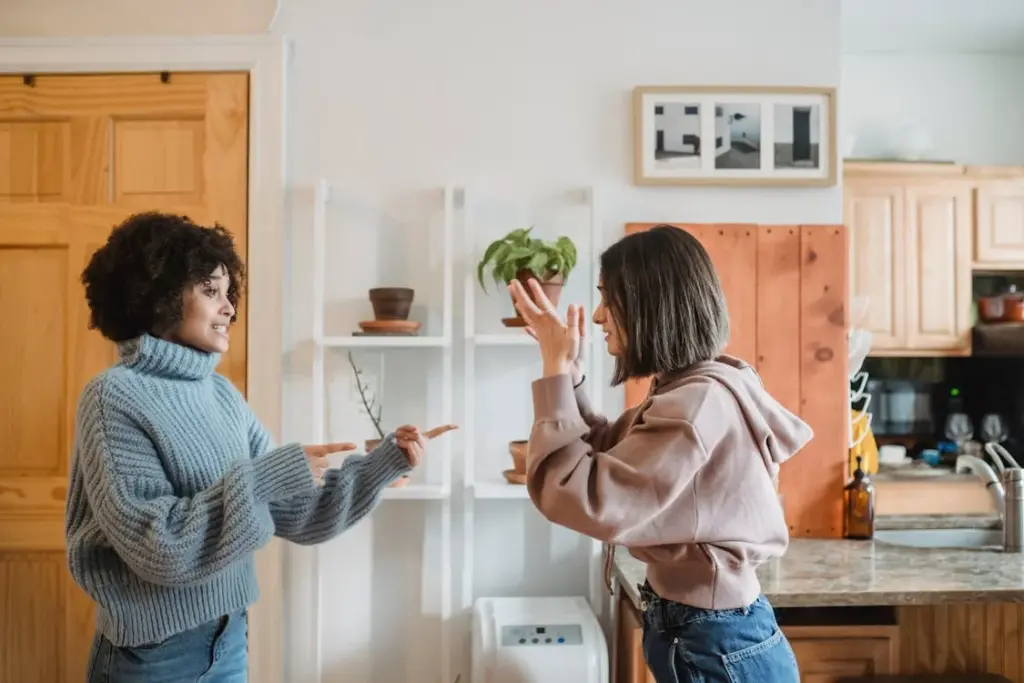 Two women in casual clothing are engaged in conversation in a cozy kitchen with wooden cabinets and indoor plants.