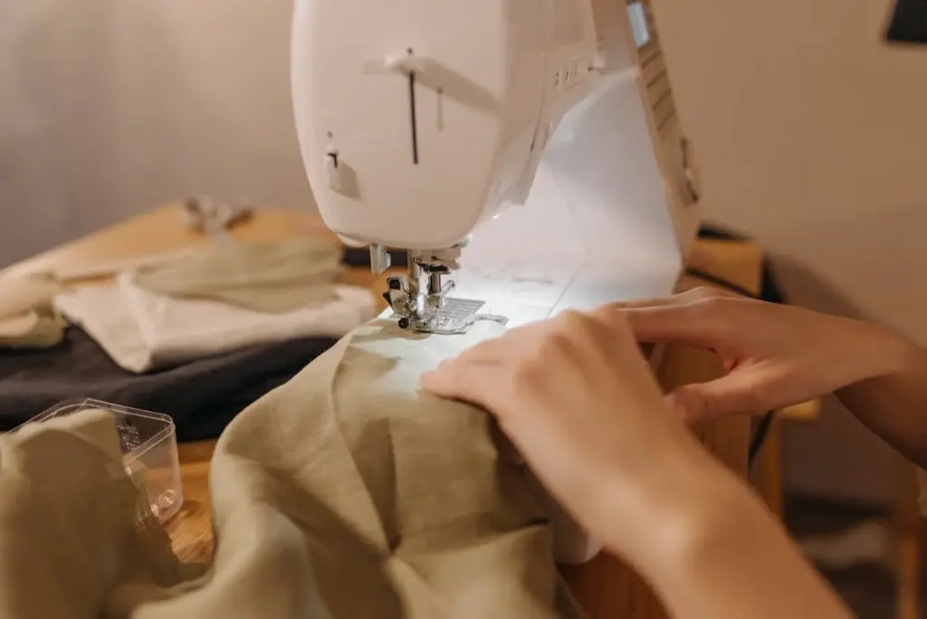 A sewing machine being used to stitch green fabric, with hands guiding the material. Other fabric pieces and sewing tools are visible on the wooden table nearby.
