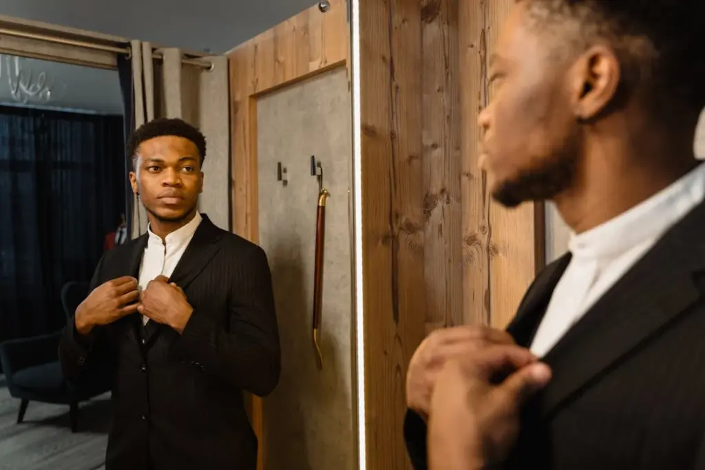 A man in a suit looks at himself in a mirror while adjusting his jacket in a room with wooden walls.