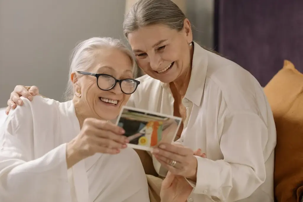 Two older women smiling while looking at a photograph together, sitting on a couch with one arm around each other.