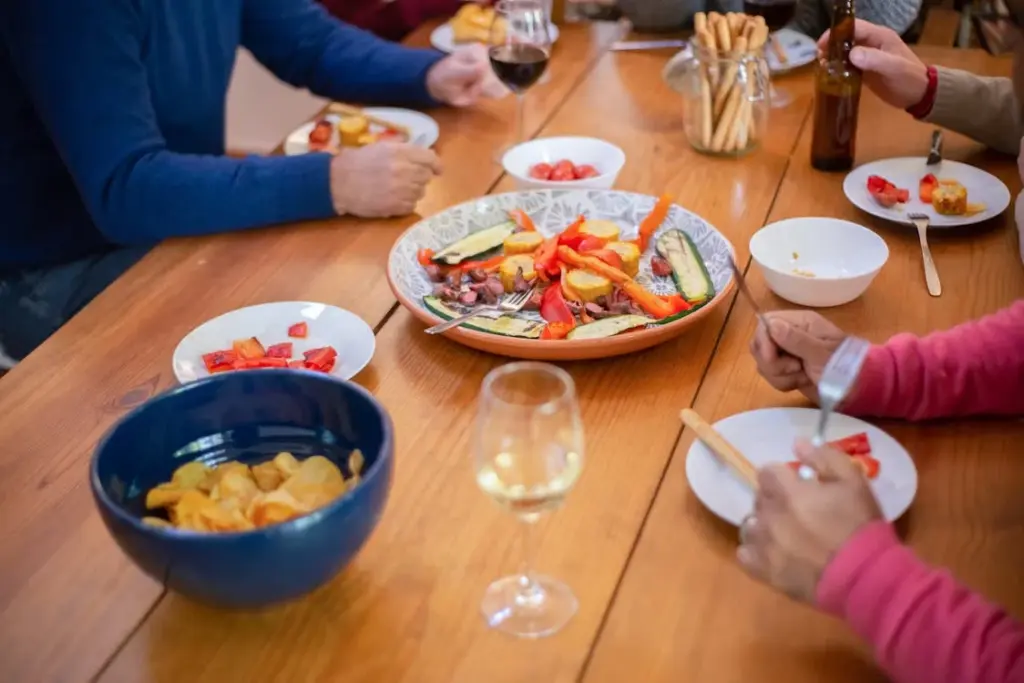 A group of people sitting around a wooden table with various dishes, including a large bowl of salad, chips, and beverages.