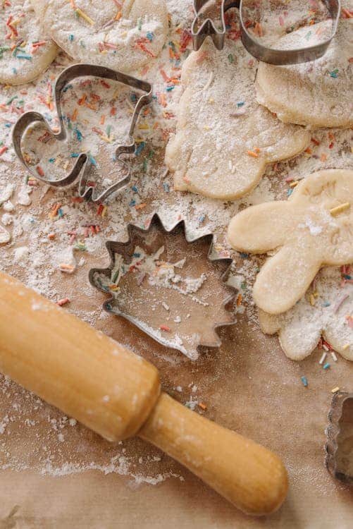 A rolling pin on a table with cookies, dough, and cookie cutters in various shapes, dusted with flour and sprinkles.