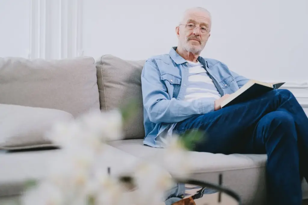 An older man with white hair and a beard sits on a gray couch, wearing a blue denim jacket and jeans, holding an open book.