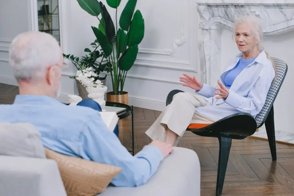 A man and a woman are engaged in a conversation while seated in a living room with white walls, plants, and a marble fireplace.