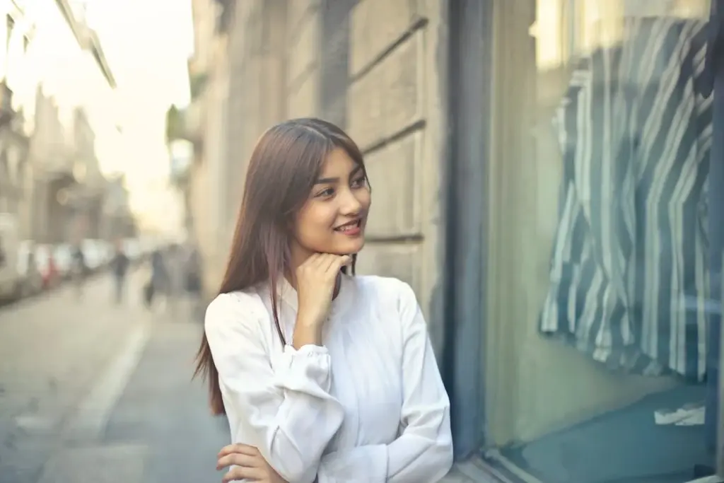 A person with long hair in a white shirt smiles while looking at a window display on a city street.