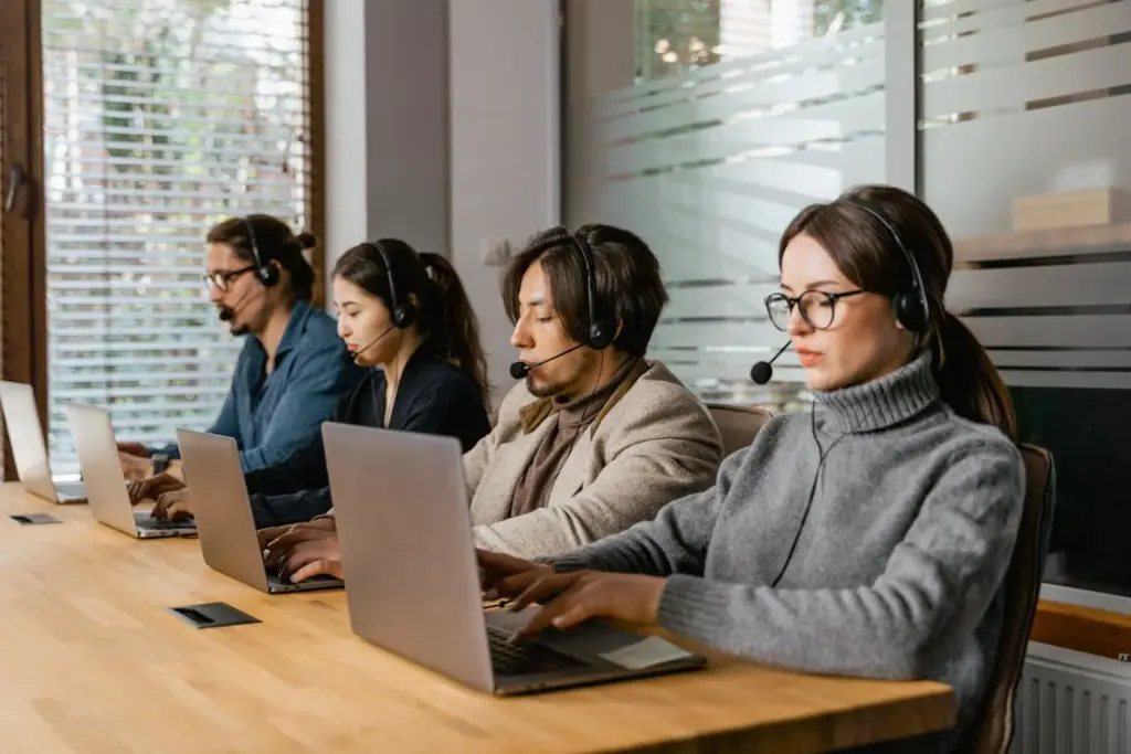Four people sitting in a row at a wooden table, wearing headsets and working on laptops.