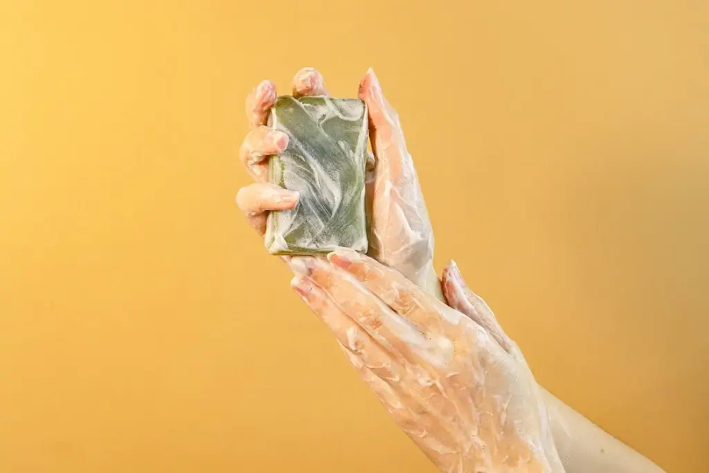 Hands covered in soap lather holding a gray soap bar against a yellow background.