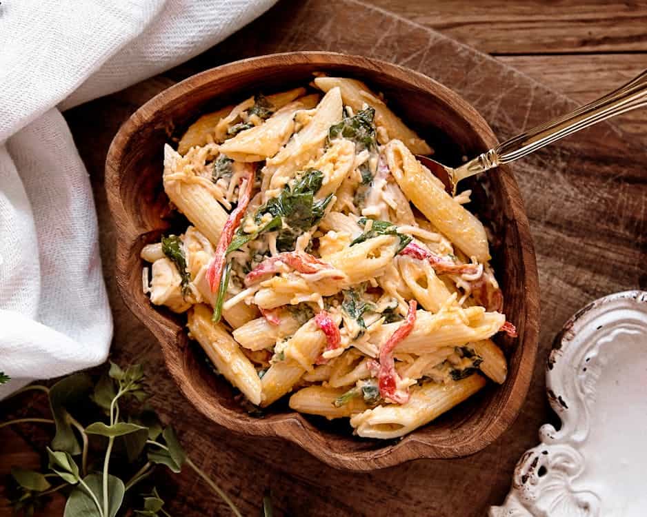 A wooden bowl filled with creamy pasta mixed with spinach and red bell peppers on a wooden surface with a white cloth and herbs nearby.