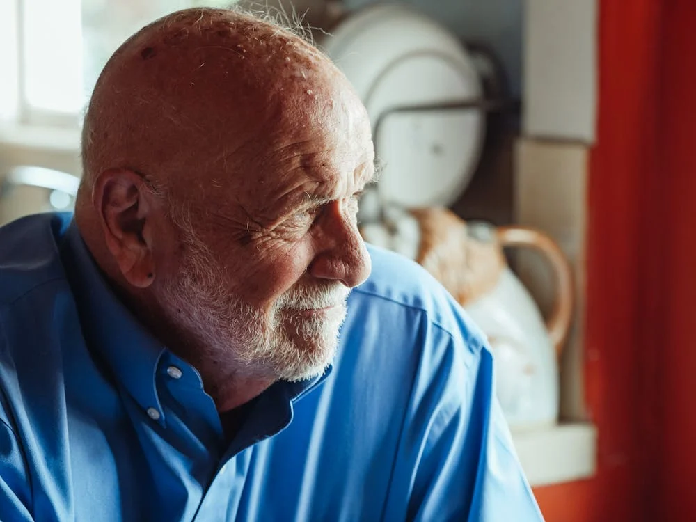 An older man with a bald head and white beard, wearing a blue shirt, looks to the side in a brightly lit room with dishes and kitchenware in the background.