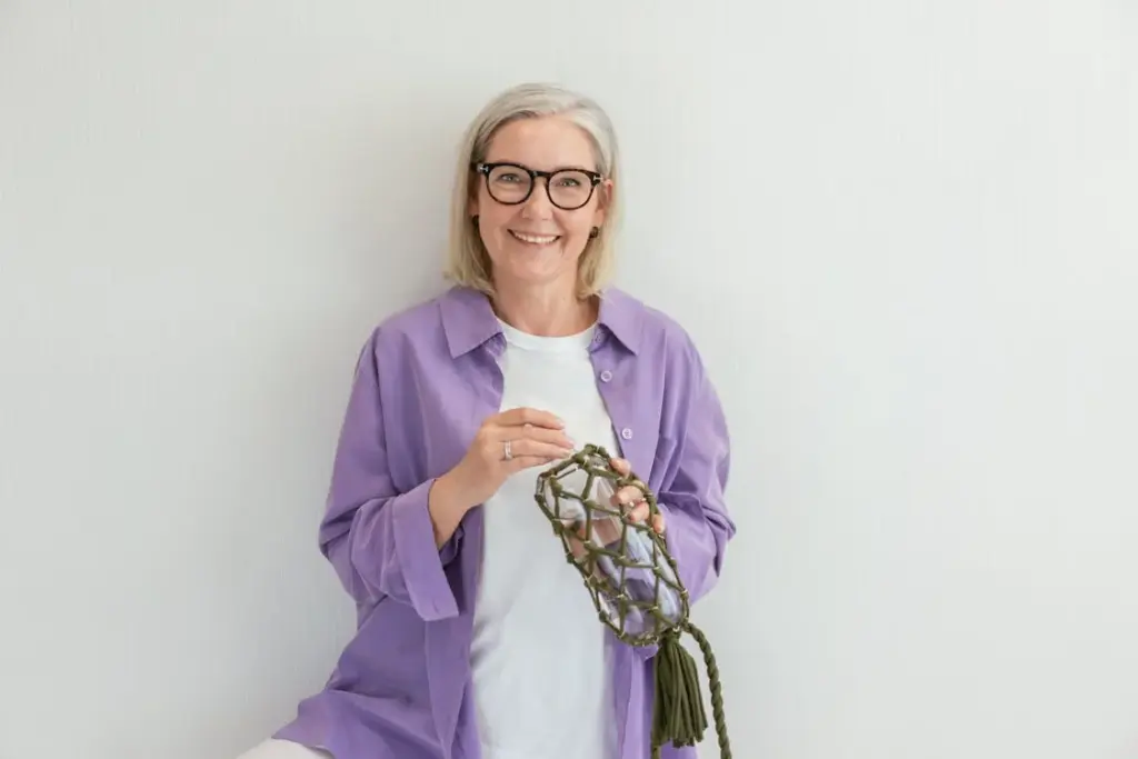 A smiling person with short gray hair and glasses is holding a green knotted rope object. They are wearing a light purple shirt and standing against a plain white background.