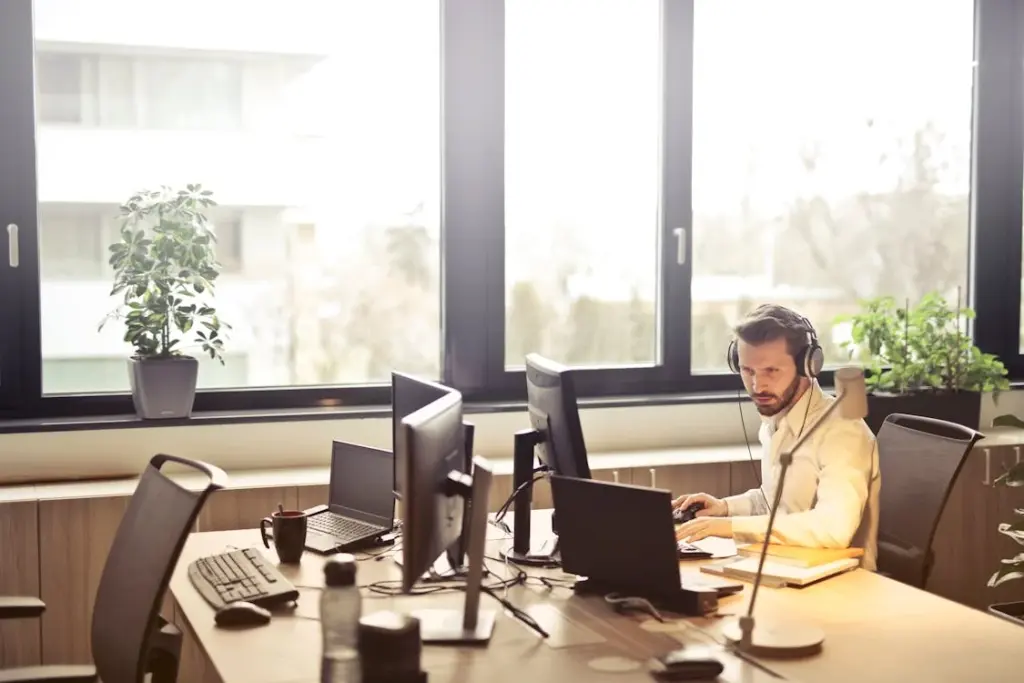 A person wearing headphones works on multiple computer screens in a bright office with large windows and potted plants.