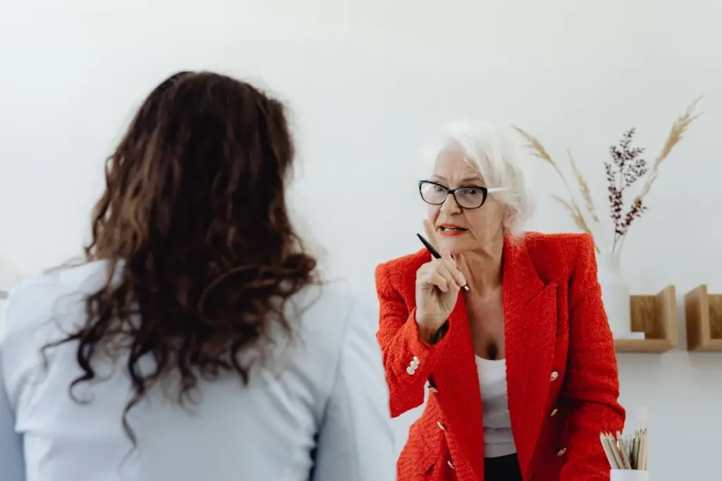 An older woman in a red blazer is speaking to a person with long hair facing away from the camera, gesturing with a pen in a well-lit room.