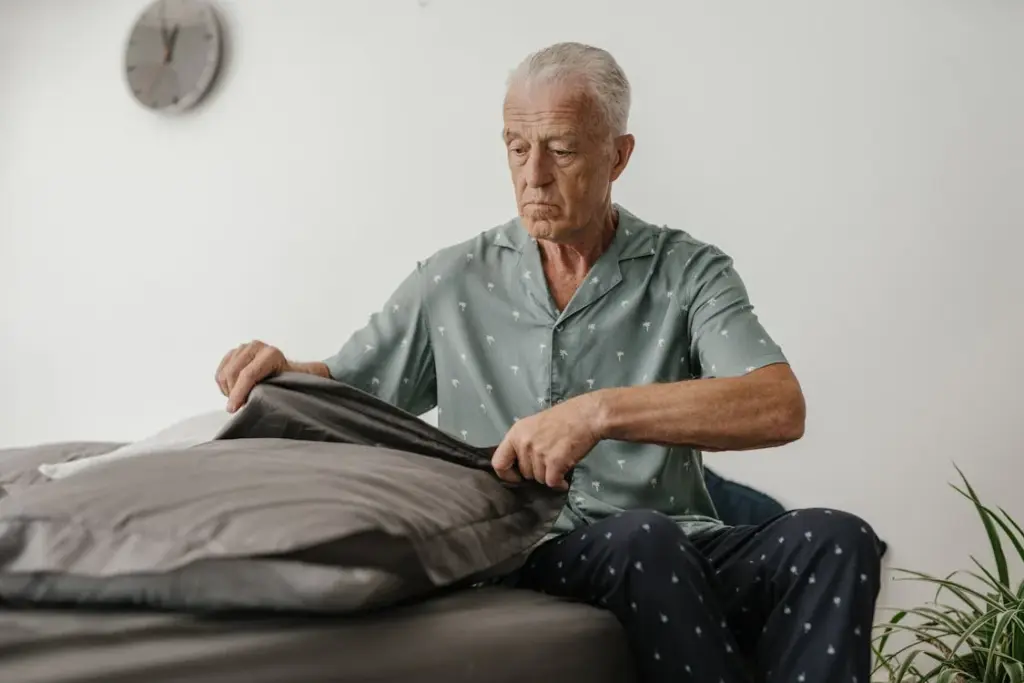 An elderly man in pajamas sits on a bed, tucking in gray bedding. A clock is visible on the wall in the background.