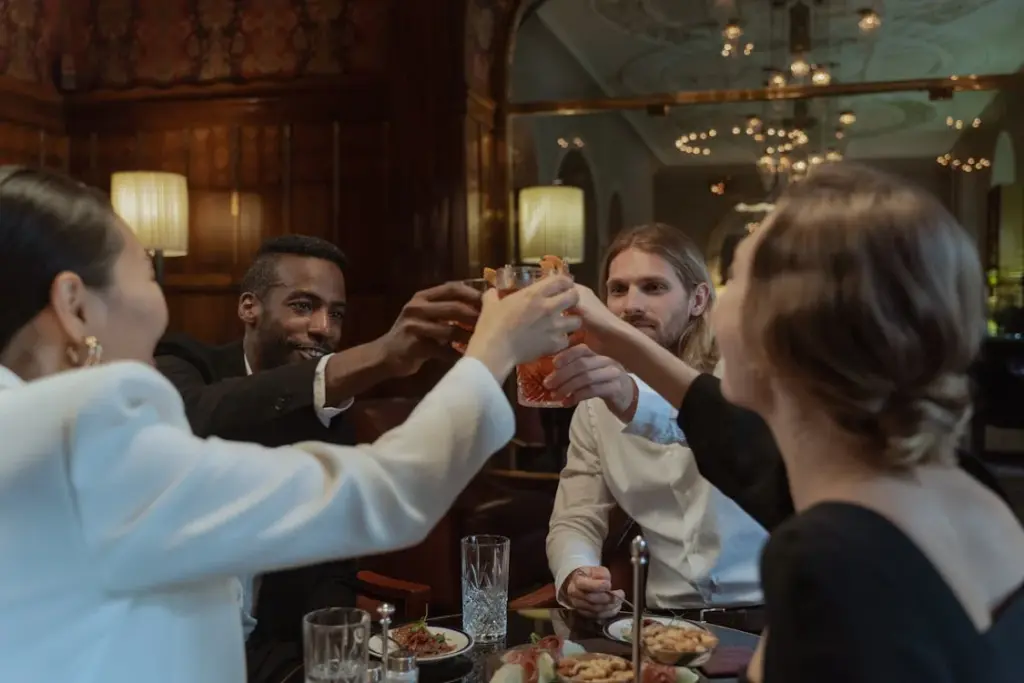 A group of four people, dressed formally, raising their glasses in a toast at a dining table in an elegant restaurant.