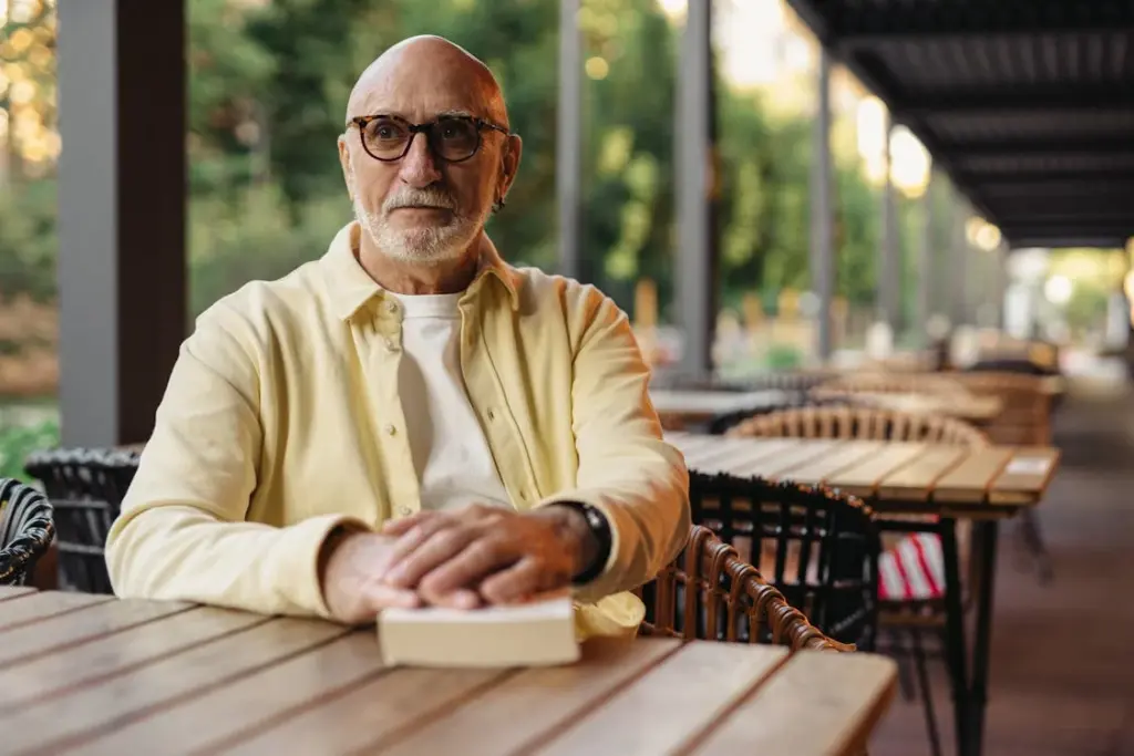 A man with glasses and a yellow shirt sits at an outdoor cafe table with a book.