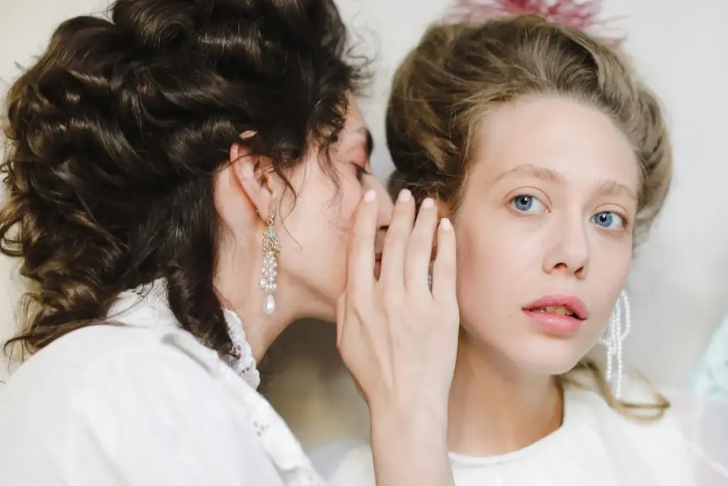 Two older women, dressed in period attire and elaborate hairstyles, share a secret. One whispers into the other's ear, who looks forward with a calm expression. Both are adorned with pearl earrings, their age making the whispered exchange seem less important.