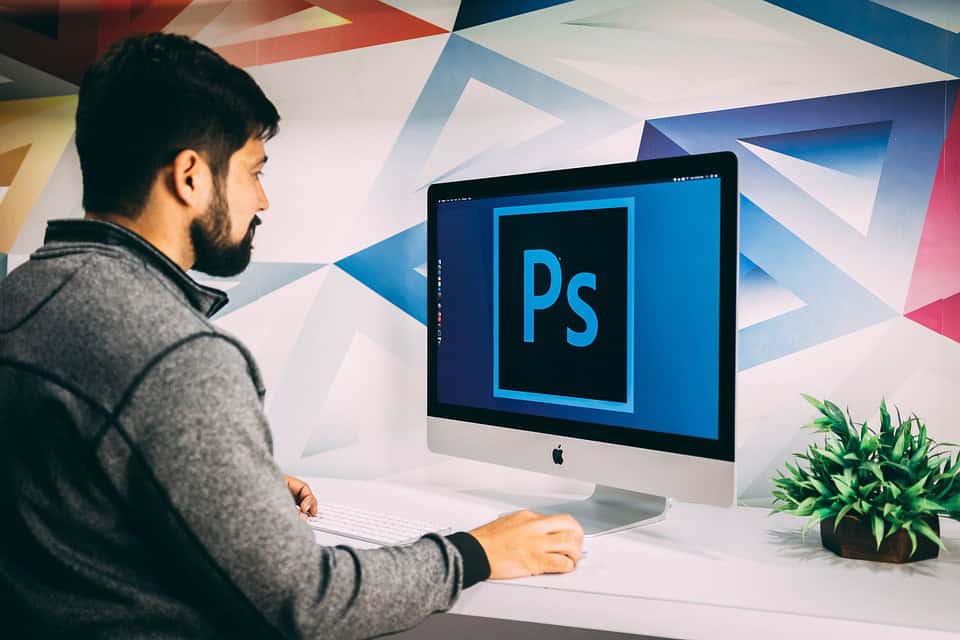 A man sits at a desk and works on a computer displaying the Adobe Photoshop logo.
