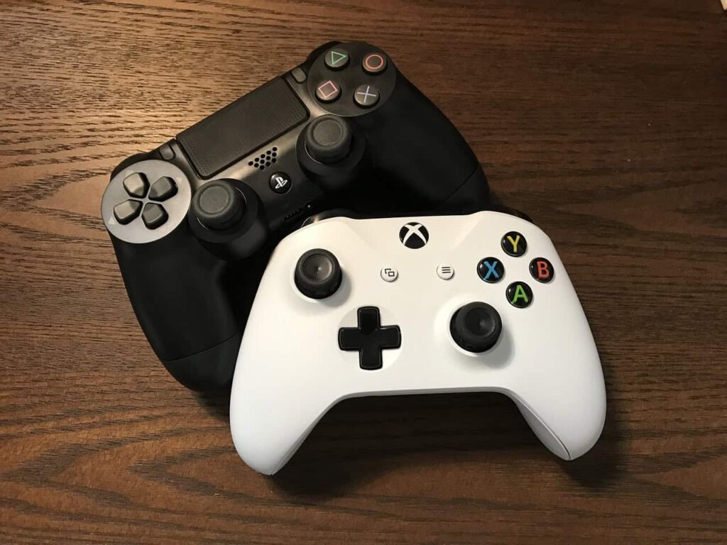 A black PlayStation controller and a white Xbox controller are placed on a wooden surface.
