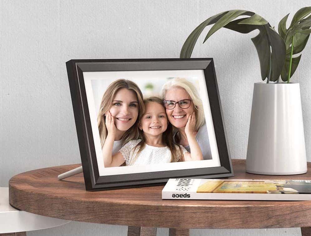 A framed photo of three women, likely from different generations, is placed on a round wooden table next to a white vase with green leaves and a book.