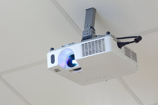 Ceiling-mounted projector with a blue lens light, attached to a white ceiling tile.