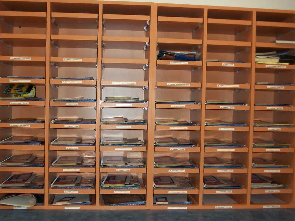 Wooden shelves with numerous compartments, each labeled with a name. Various documents and folders are placed in some compartments, while others are empty.