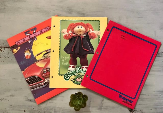 Three vintage folders: one with a '57 Heaven car design, one featuring a Cabbage Patch Kid, and a red Trapper Keeper, laid out on a wooden surface with a small green plant.