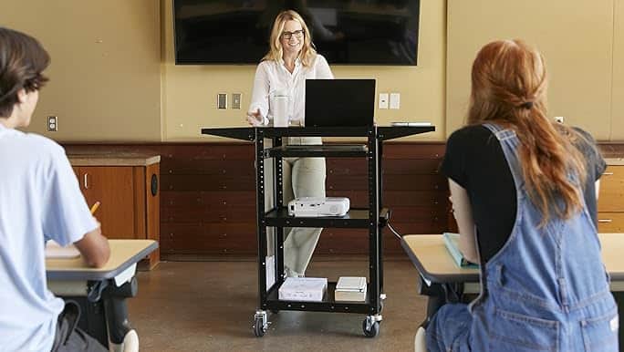A woman stands behind a laptop on a black mobile cart, addressing two seated individuals in a room with a wall-mounted TV.