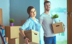 Two people carrying boxes and a plant, standing in a room with additional cardboard boxes and unpacked items.