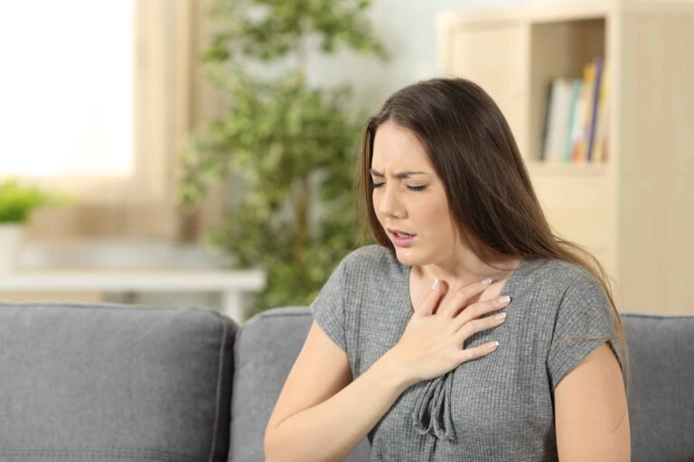 A woman sitting on a couch with one hand on her chest, appearing to be in pain or discomfort.