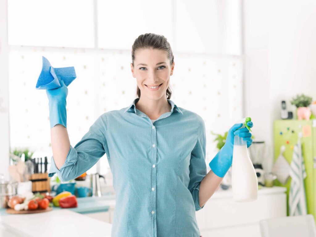 A woman wearing blue gloves is holding a blue cloth in one hand and a spray bottle in the other, standing in a kitchen.