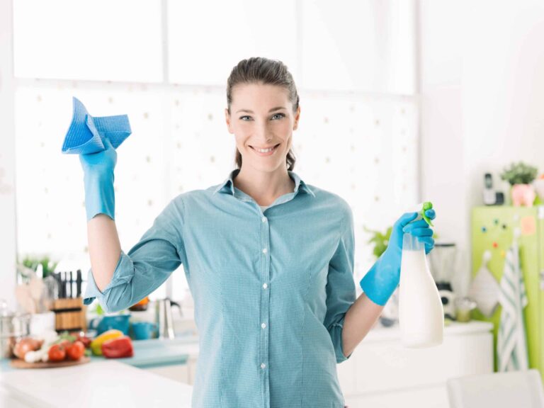 A woman wearing blue gloves is holding a blue cloth in one hand and a spray bottle in the other, standing in a kitchen.