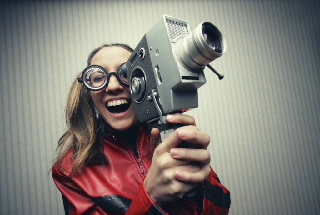 A person with large round glasses, wearing a red jacket, smiles widely while holding and aiming an old-fashioned film camera.