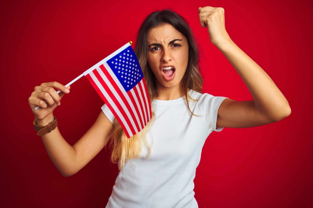 Woman in a white t-shirt passionately holding and raising a small American flag, with her other hand clenched in a fist, against a red background.