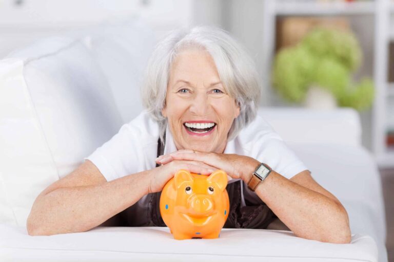 Elderly woman with white hair smiles while resting her hands on an orange piggy bank on a white couch.