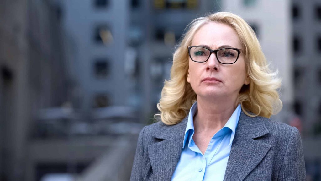 A woman with blonde hair and glasses, wearing a gray blazer and blue shirt, stands outdoors in an urban setting looking serious.