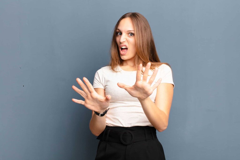 A woman with long brown hair, wearing a white shirt and black pants, stands against a gray background with her hands raised in a defensive gesture and an expressive look on her face.