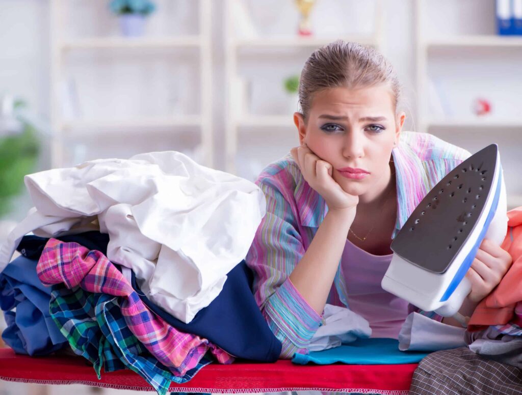 A person with a bored expression holds an iron while leaning on a pile of clothes on an ironing board.