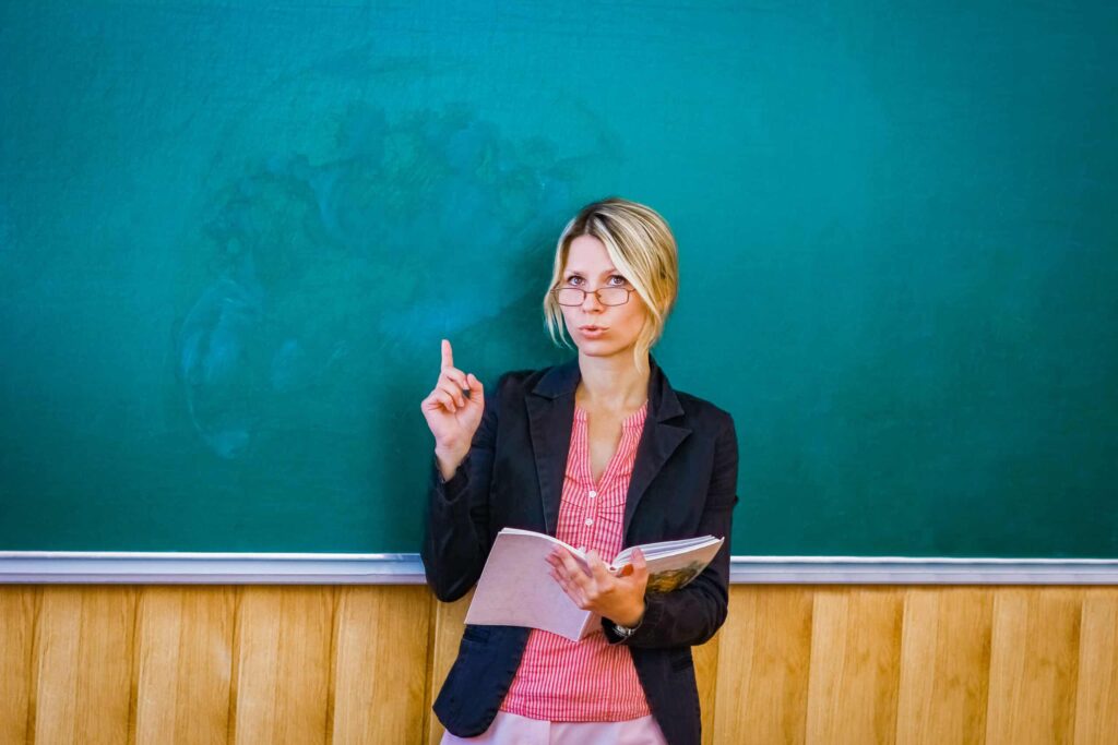A person wearing glasses, holding a book, and pointing upward stands in front of a green chalkboard.