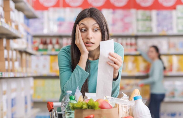 A woman in a teal shirt looks surprised while holding a long receipt in a grocery store. A filled shopping cart is in front of her, and another shopper is seen in the background.