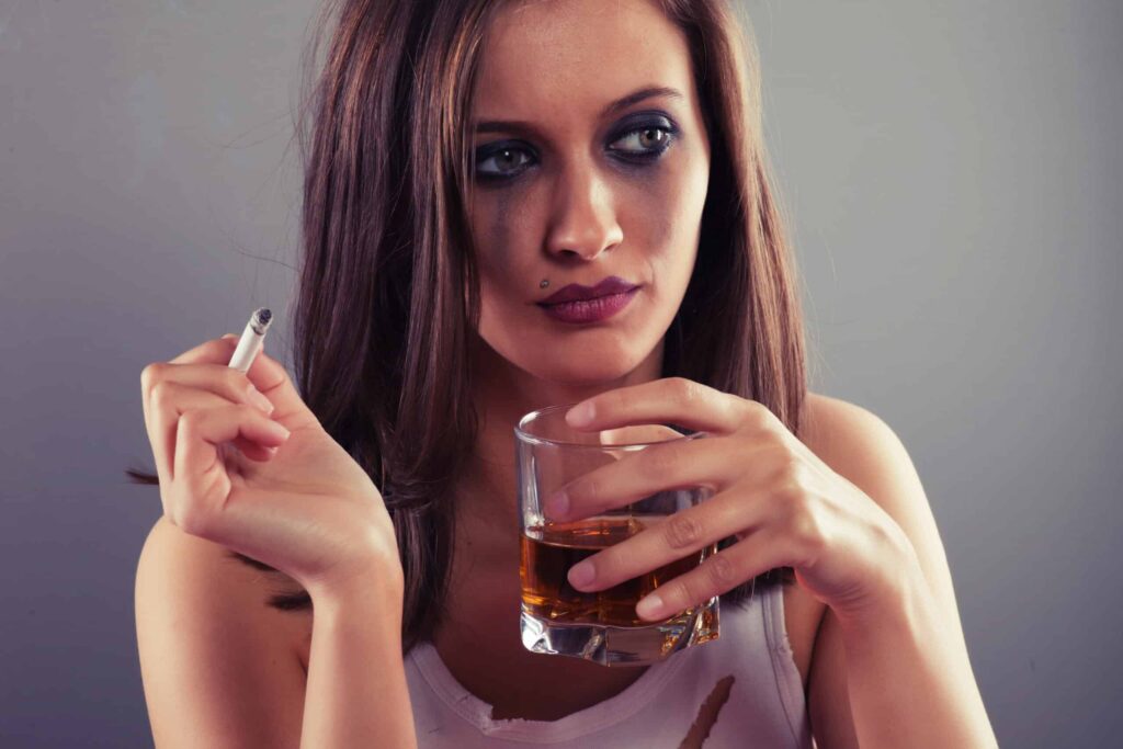 A woman with smeared makeup holds a cigarette in her left hand and a glass of whiskey in her right hand, looking contemplative.