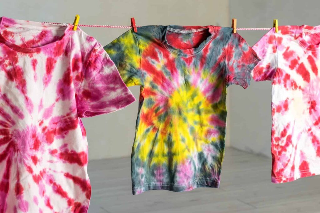 Three tie-dye t-shirts in red, yellow, green, and pink hues are hanging on a clothesline with clothespins.