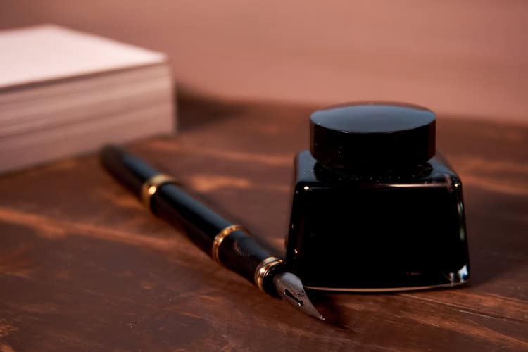 A black fountain pen and an ink bottle are placed on a wooden desk next to a stack of white paper sheets.
