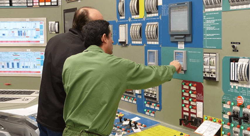 Two men in a control room, with one man in a green uniform pointing at a control panel while the other observes. Various dials, switches, and monitors are mounted on the wall.