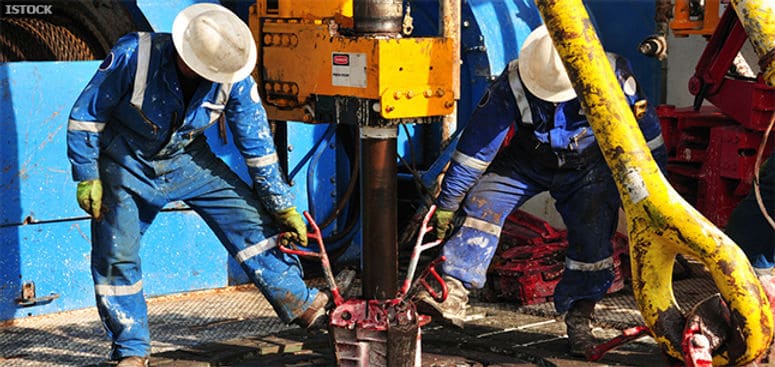 Two oil rig workers in blue coveralls and hard hats operate heavy drilling machinery on a platform.