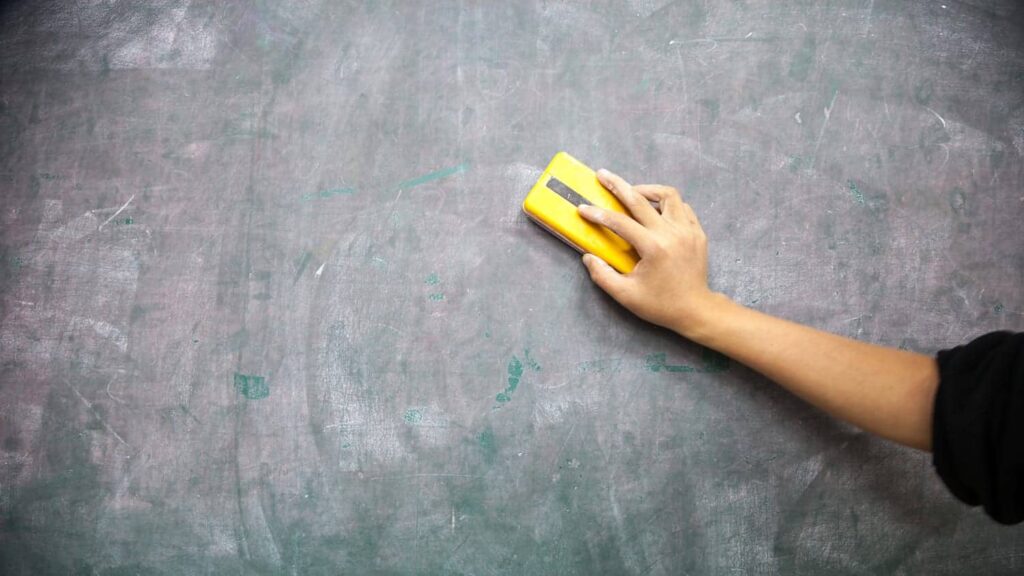 A hand erases chalk markings from a green chalkboard using a yellow eraser.
