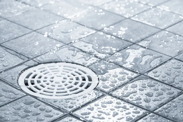 Close-up of a wet tiled floor with a white round drain cover. Water droplets are scattered across the dark gray tiles.