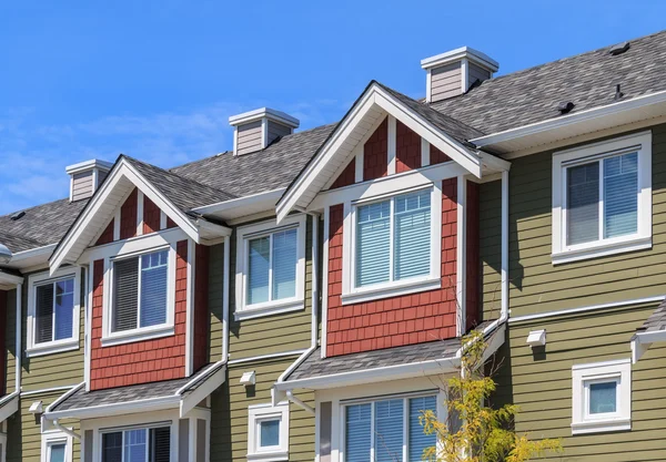 A row of modern townhouses with green and red facades featuring gable roofs, white trim, and large windows under a clear blue sky.