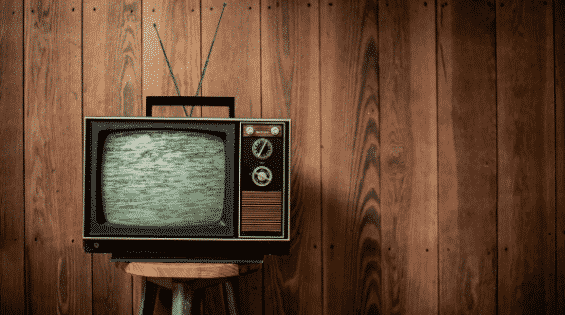 A vintage television with antennas stands on a stool in front of a wooden wall. The screen shows static.