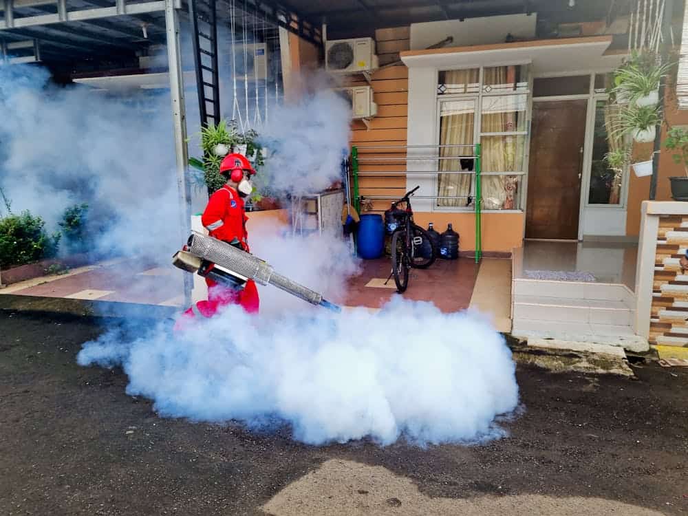 Person in red protective gear using a fogging machine to disperse smoke or insecticide outside a residential house, with a bicycle and potted plants nearby.