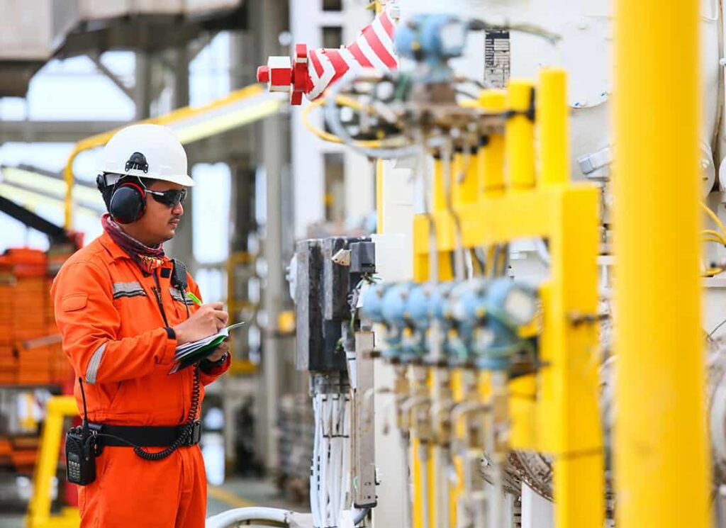 A worker in an orange jumpsuit and hard hat inspects equipment at an industrial site, using a notebook and wearing safety gear, including ear protection and sunglasses.